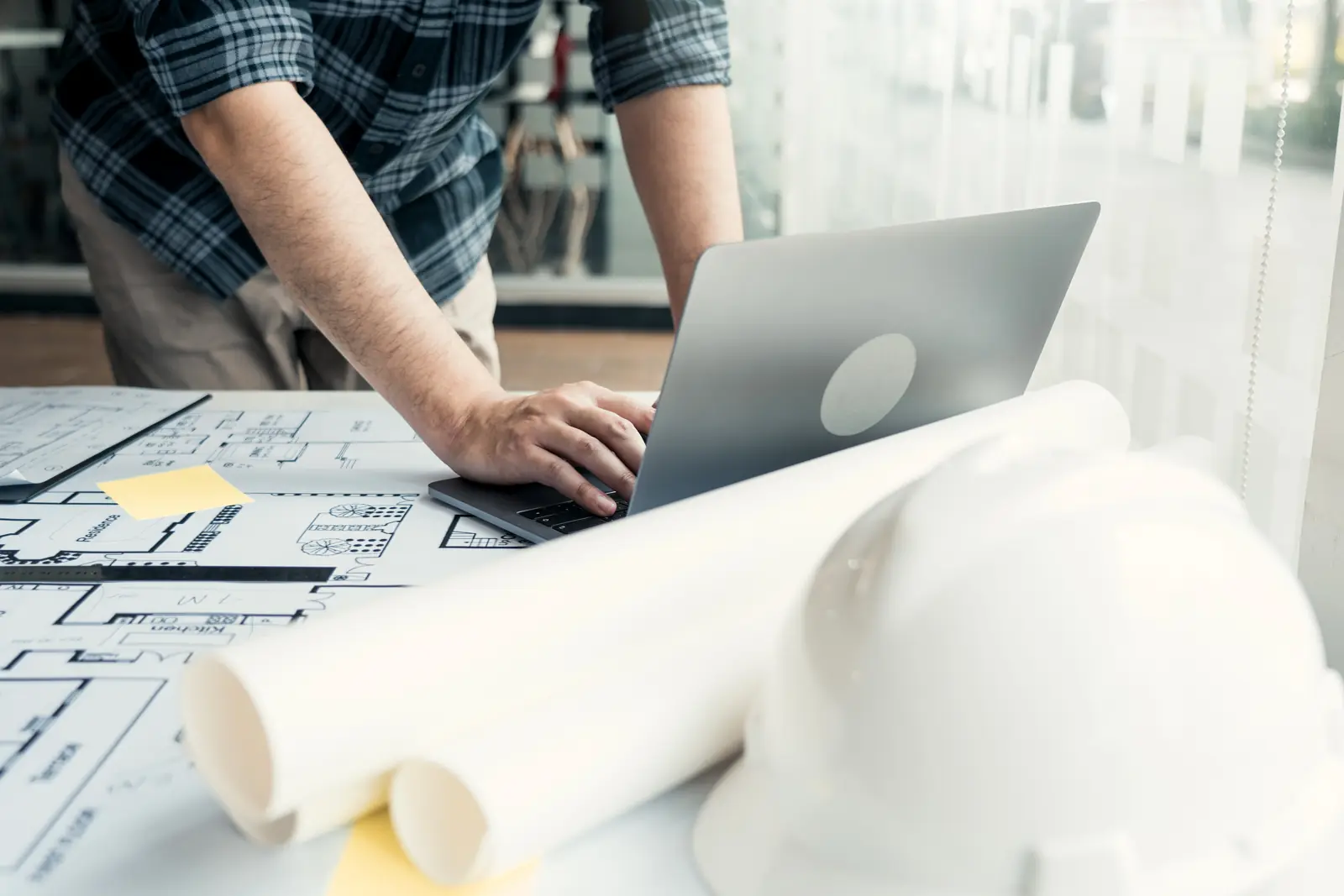 Estimator working on a laptop over rolled construction plans and a hard hat