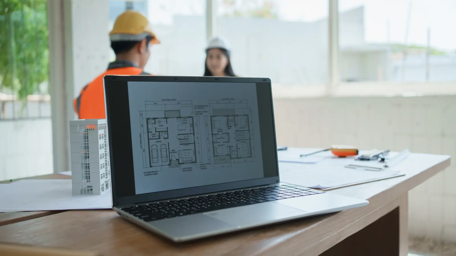 Laptop showing floor plans on a desk in a construction site office, two workers in hard hats behind it